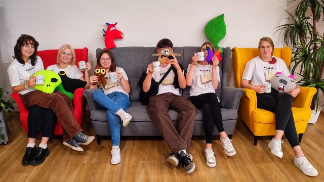 młodzież siedzi na sofie i fotelach i prezentuje uszyte prze siebie maskotki