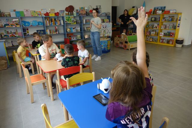 dzieci siedzą przy stolikach, przed nimi stoi kobieta, w tle regały kolorowe, na stolikach roboty Photon