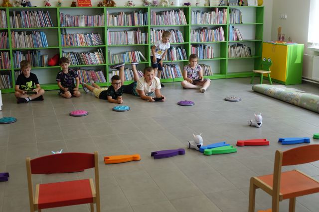 dzieci sterują robotami jeżdżącyni po torze przeszkód, w tle regały z książkami