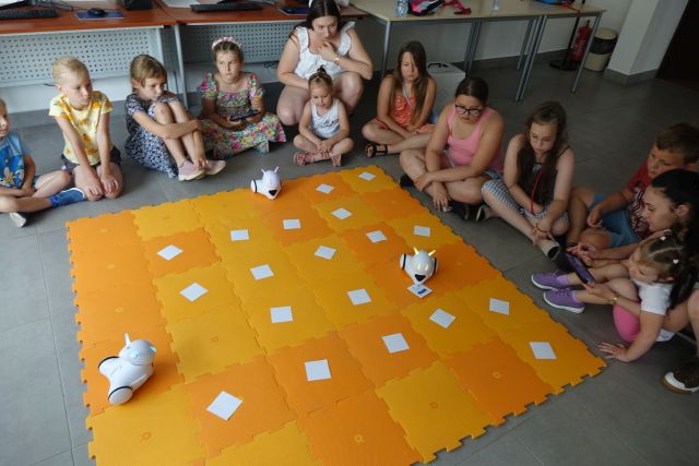 dzieci sterują przy użyciu tabletów robotami jeżdżącymi po pomarańczowej macie