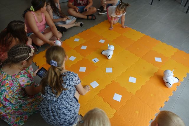 dzieci sterują przy użyciu tabletów robotami jeżdżącymi po pomarańczowej macie