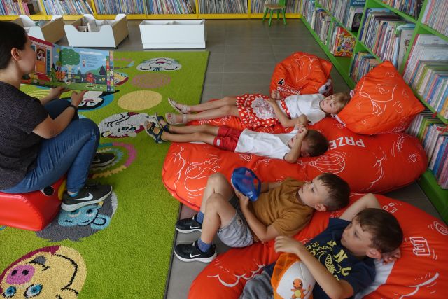 dziewczynka i 3 chłopców leżą na czerwonych pufach, przed nimi kobieta siedzi i czyta książkę