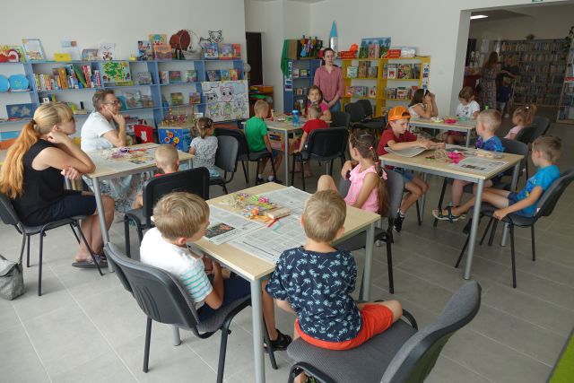 dzieci siedzą przy stolikach w sali bibliotecznej, przed nimi stoi kobieta