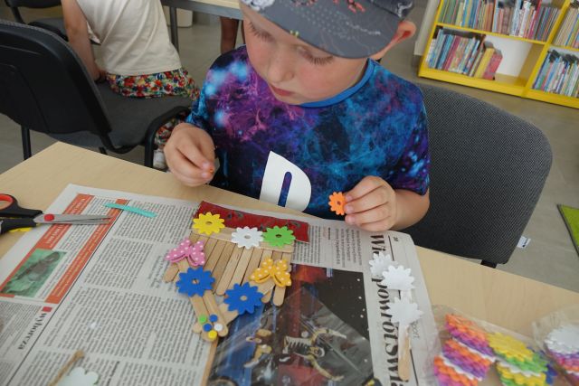 chłopiec robi z patyczków domek i nakleja na niego kwiatki z materiałów