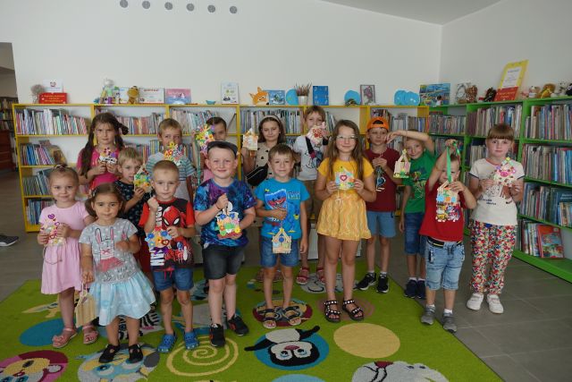 grupa około 15 dzieci pozuje do zdjęcia z wykonanymi przez siebie domkami z patyczków wyklejonymi ozdobami z materiałów, w tle regały z książkami