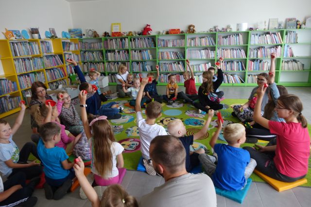 dzieci i rodzice siedzą w okręgu na kolorowym dywanie, w tle regały z książkami