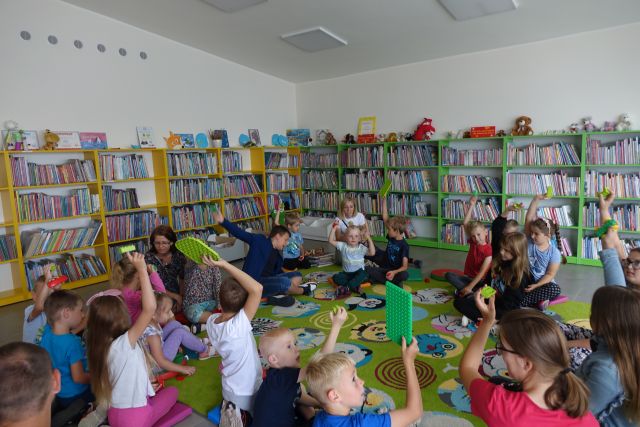 dzieci siedzą na zielonym dywanie i trzymają w podniesionych dłoniach kolorowe klocki