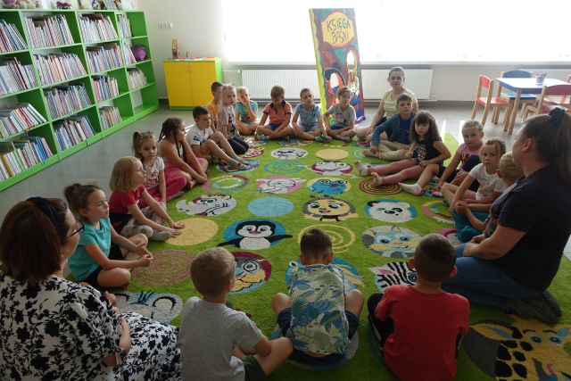 dzieci siedzą w okręgu na zielonym dywanie, w tle regały z książkami