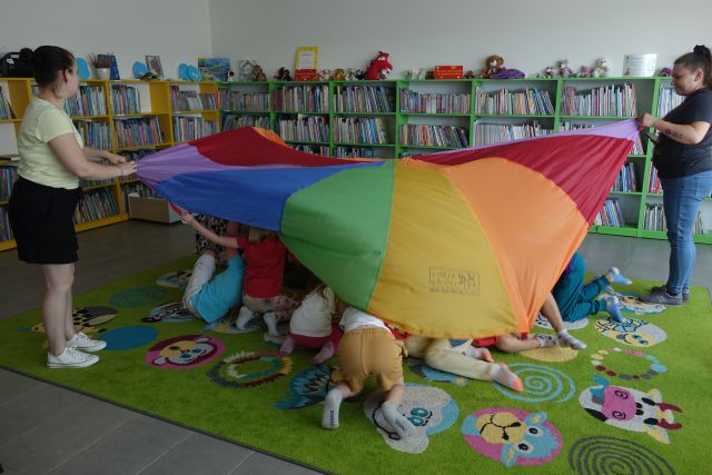 trzy kobiety trzymają kolorową chustę animacyjną, a pod nią chowają się dzieci