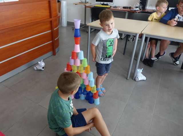 uczestnicy zajęć programują układając według kodu kolorowe kubeczki, w tle małe białe roboty