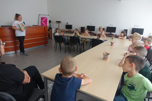 przedszkolaki siedzą przy stołąch ustawionych w podkowę, przed nimi stoi bibliotekarka z książką