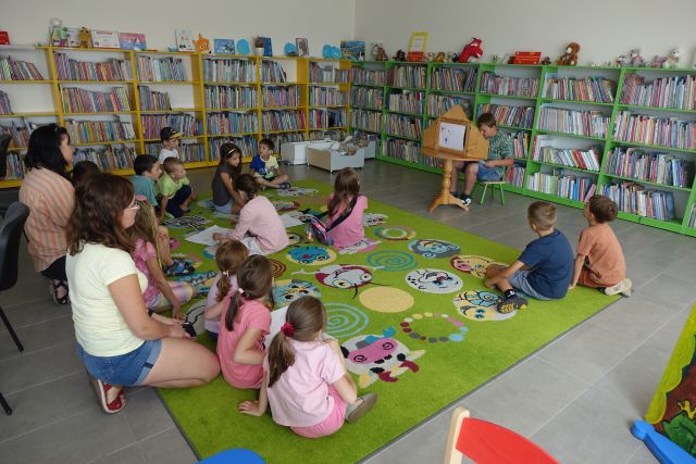 dzieci i rodzice siedzą na kolorowym dywanie, przed nimi chłopiec pokazuje ilusrracje w teatrzyku kamishibai