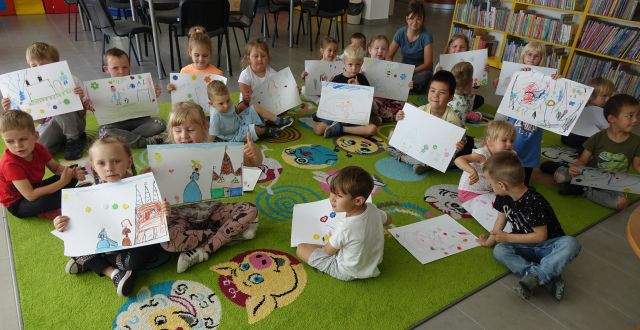 przedszkolaki siedzące na kolorowym dywanie i prezentujące swoje ilustracje