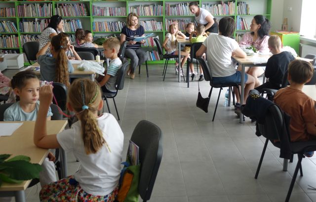 mali i duzi uczestnicy warsztatów siedzą przy stolikach, przed nimi siedzi kobieta i czyta książkę, w tle regały z książkami