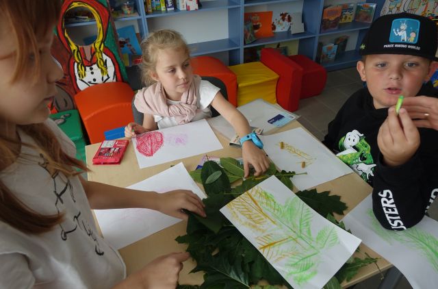 na stokiu kartki, kredki, liście, przy stoliku siedzą dzieci