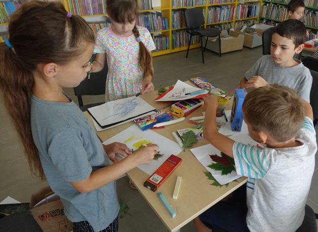 na stokiu kartki, kredki, liście, przy stoliku stoją dzieci i malują
