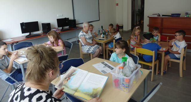 uczestnicy warsztatów mali i duzi siedzą przy stołąch i słuchają czytanej przez bibliotekarkę bajki