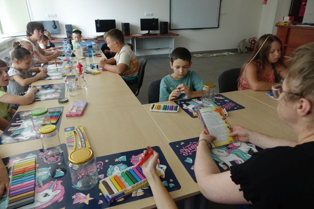 dzieci siedzą przy stołąch i słuchają jak bibliotekarka czyta książkę