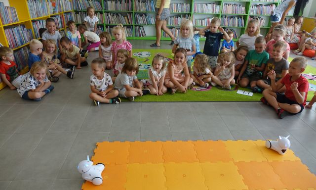 przedszkolaki siedzą na kolorowym dywanie, przed nimi na pomarańczowej macie roboty Photon, w tle regały z książkami