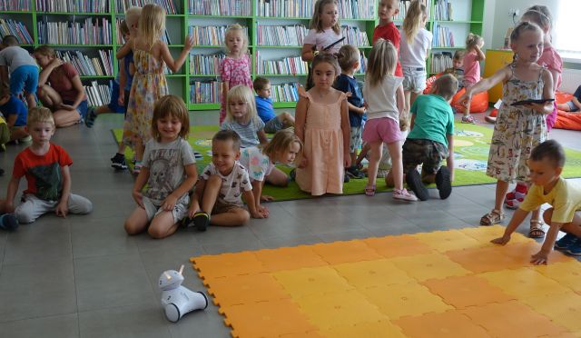 przedszkolaki siedzą na kolorowym dywanie, przed nimi na pomarańczowej macie roboty Photon, w tle regały z książkami