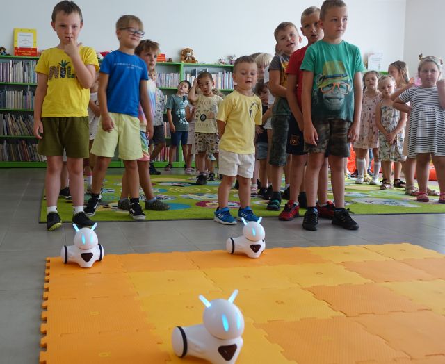 przedszkolaki stoją na kolorowym dywanie, przed nimi na pomarańczowej macie roboty Photon, w tle regały z książkami
