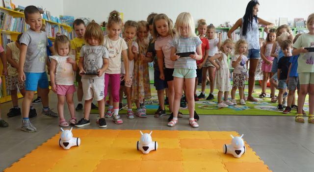 przedszkolaki stoją na kolorowym dywanie, sterują przy pomocy tabletów robotami Photon, w tle regały z książkami