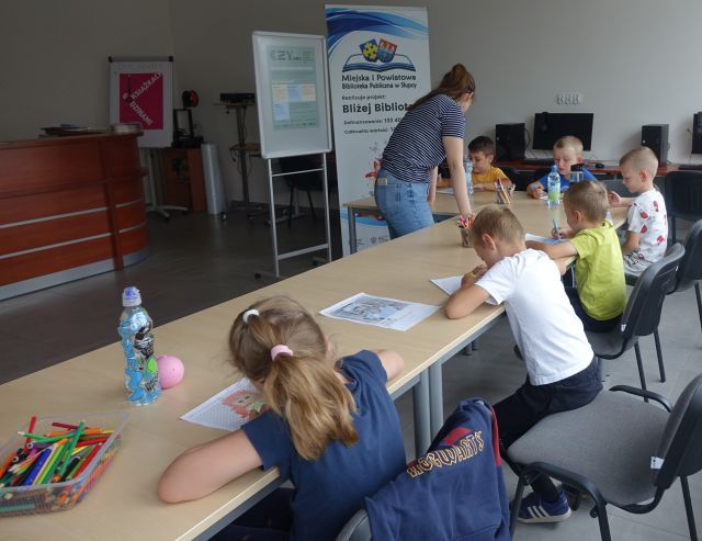 bibliotekarka pomaga dzieciom siedzącym przy stolikach wypełniać krzyżówki