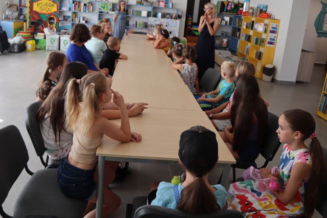 20 dzieci siedzi przy długim stole, przed nimi stoją dwie kobiety