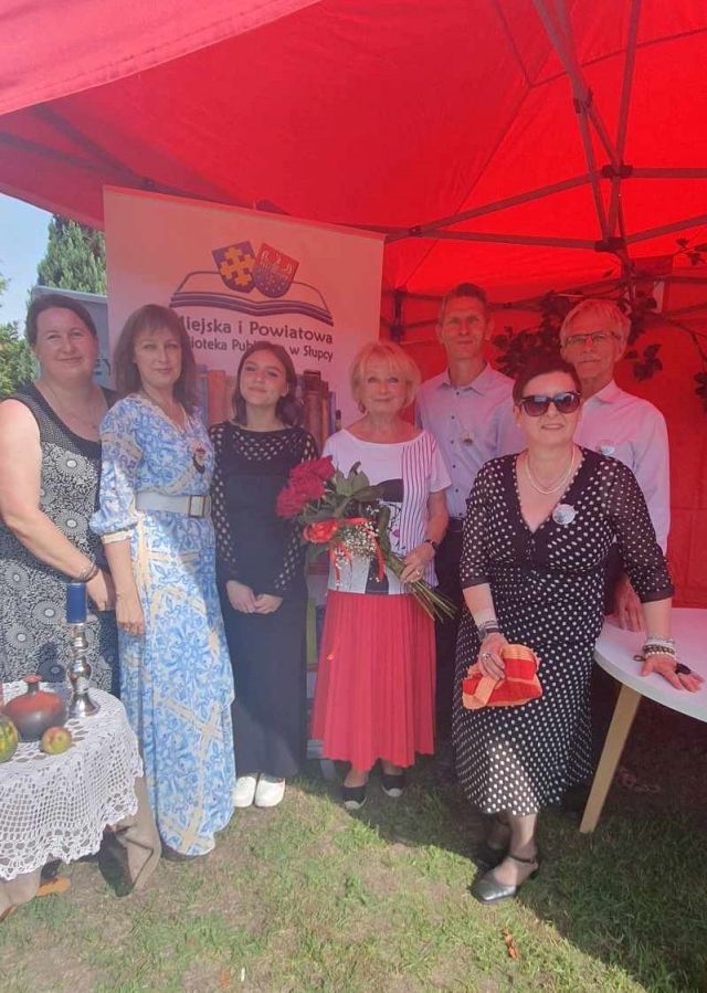 do zdjęcia pozują od lewej strony kobieta w ciemnej sukience, kobieta w niebieskiej sukience, dziewczyna w czarnej sukience, aktorka w białęj bluzce, czerwonej spódnicy i czerwonymi różami w dłoniach, dwóch mężczyzn oraz przed nimi kobieta w sukience w czarne grochy