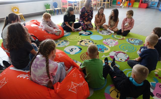 dzieci uczestniczące w warsztatach siedzą na kolorowym dywanie i pomarańczowych pufach