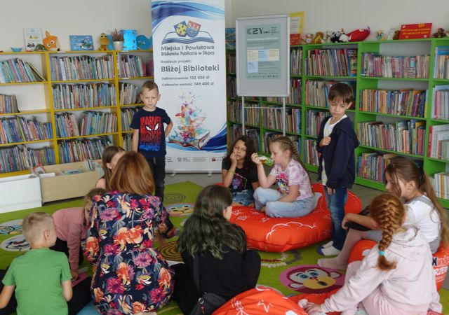 dwóch chłopców stoi a pozostałę dzieci i nauczycielka siedzą na kolorowym dywanie, w tle baner reklamujący nazwę biblioteki i projekt oraz regały z książkami