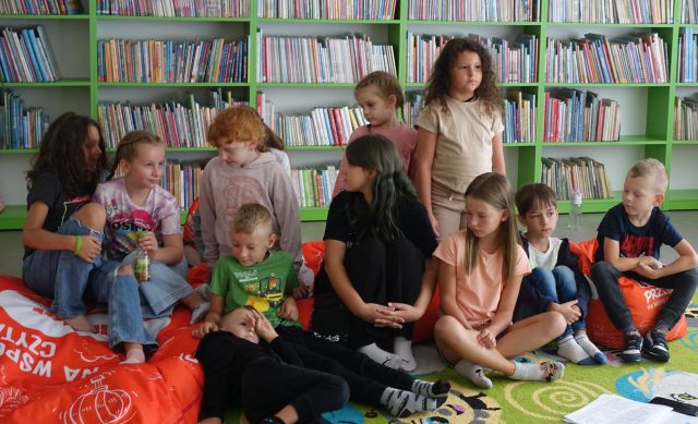 dzieci siedzą na kolorowym dywanie, w tle regały z książkami