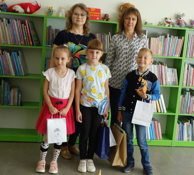 trójka dzieci z nagrodami za udział w projekcie ora dwie bibliotekarki, w tle regały z książkami