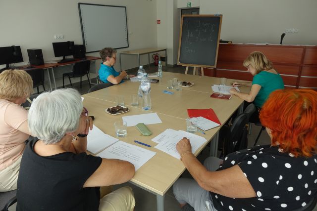5 seniorek siedzi przy stołach, przed nimi leżą kartki i długopisy
