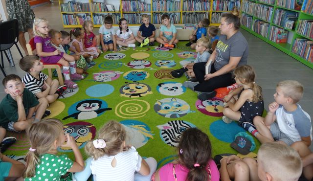 przedszkolaki siedzą w kole na kolorowym dywanie, w tle regały z książkami
