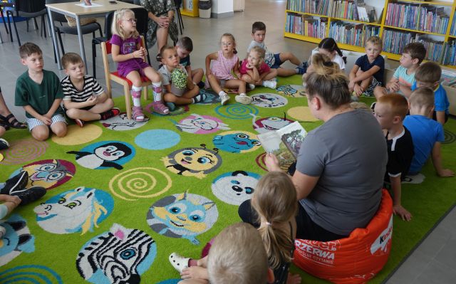 przedszkolaki siedzą na kolorowym dywanie, bibliotekarka czyta im bajkę