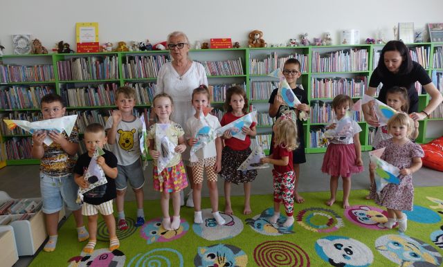 dzieci z nauczycielką i wykonanymi przez siebie ptakami z papieru pozują do zdjęcia, w tle regały z książkami