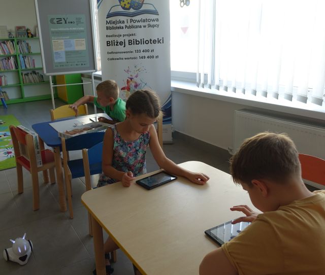 dzieci siedzą przy stolikach i sterują za pomocą tabletów robotami Photon