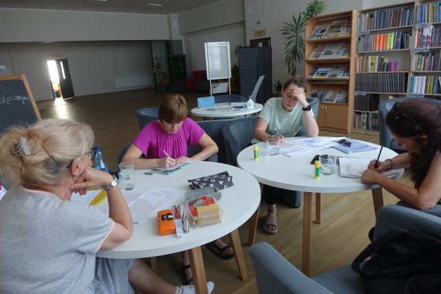 uczestniczki siedzą przy okrągłych stołąch i notują w zeszytach