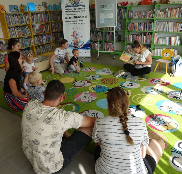 dzieci i rodzice siedzą na kolorowym dywanie i słuchają książki czytanej przez bibliotekarkę
