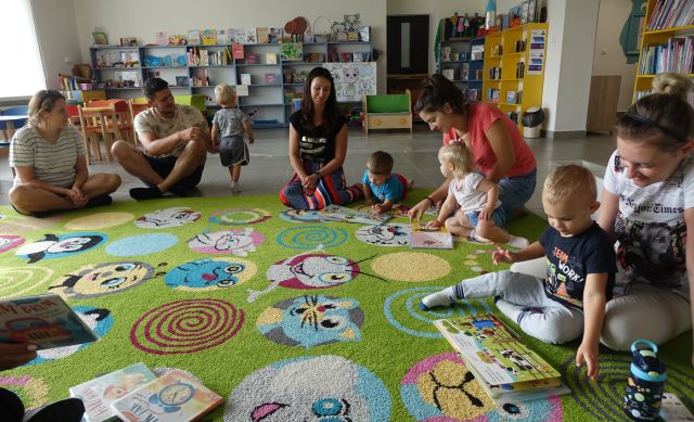 dzieci i rodzice siedzą na kolorowym dywanie i oglądają książki