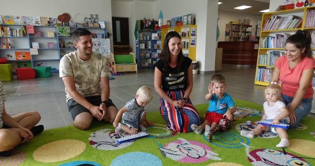 dzieci i rodzice siedzą na kolorowym dywanie, dzieci grają na instrumentach