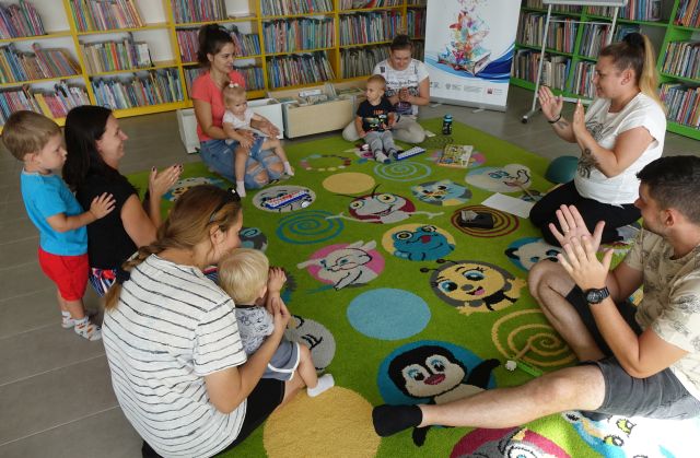 dzieci i rodzice siedzą na kolorowym dywanie i klaszczą w dłonie
