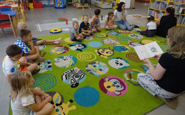 dzieci i rodzice siedzą na kolorowym dywanie, przed nimi siedzi bibliotekarka i czyta książkę