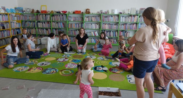 małę dzieci i rodzice siedzą na kolorowym dywanie, przed nimi kobieta trzyma książkę, w tle regały z książkami