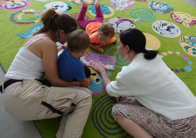 3 małych dzieci i dwie osoby dorosłe siedzą na kolorowym dywanie i czytają książkę