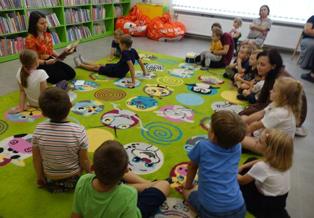 dzieci, opiekunowie i bibliotekarka siedzą na kolorowym dywanie, bibliotekarka czyta książkę, w tle regały z książkami