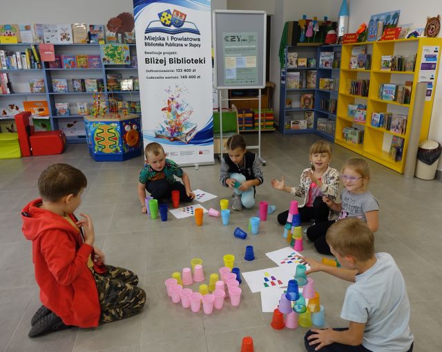 dzieci układają na podłodze wieże z kolorowych kubeczków, w tle regały i banery z nazwą projektu Bliżej Biblioteki