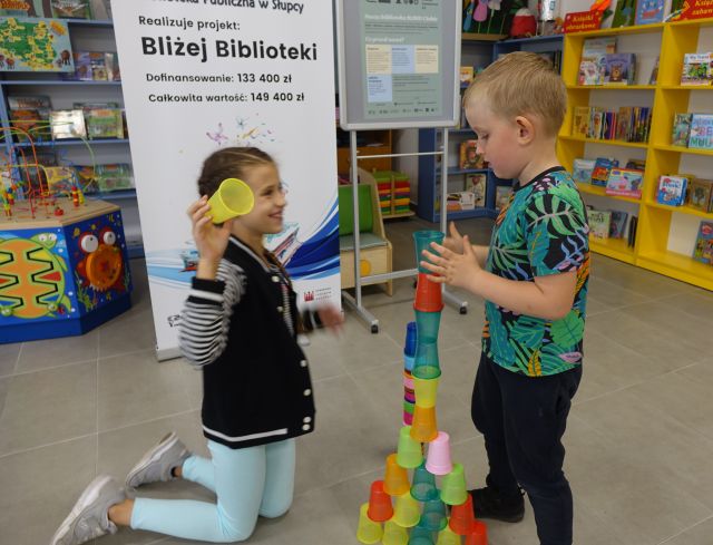chłopiec i dziewczynka ukłądają wieżę z kolorowych kubeczków, w tle banery z nazwą projektu Bliżej Biblioteki w ramach BLISKO