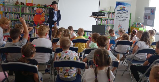 pisarz stoi przed przedszkolakami siedzącymi na krzesełkach i opowiada, w tle regały z książkami